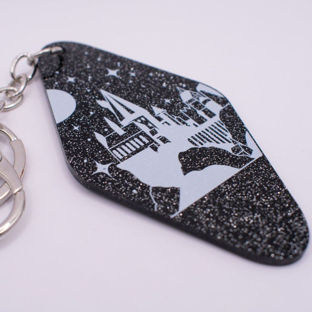 A black motel-shaped acrylic keychain with silver glitter throughout is pictured with Hogwarts castle printed on top in white. There is a silver keyring accessory with a lobster clasp attached to the top of the keyring.
