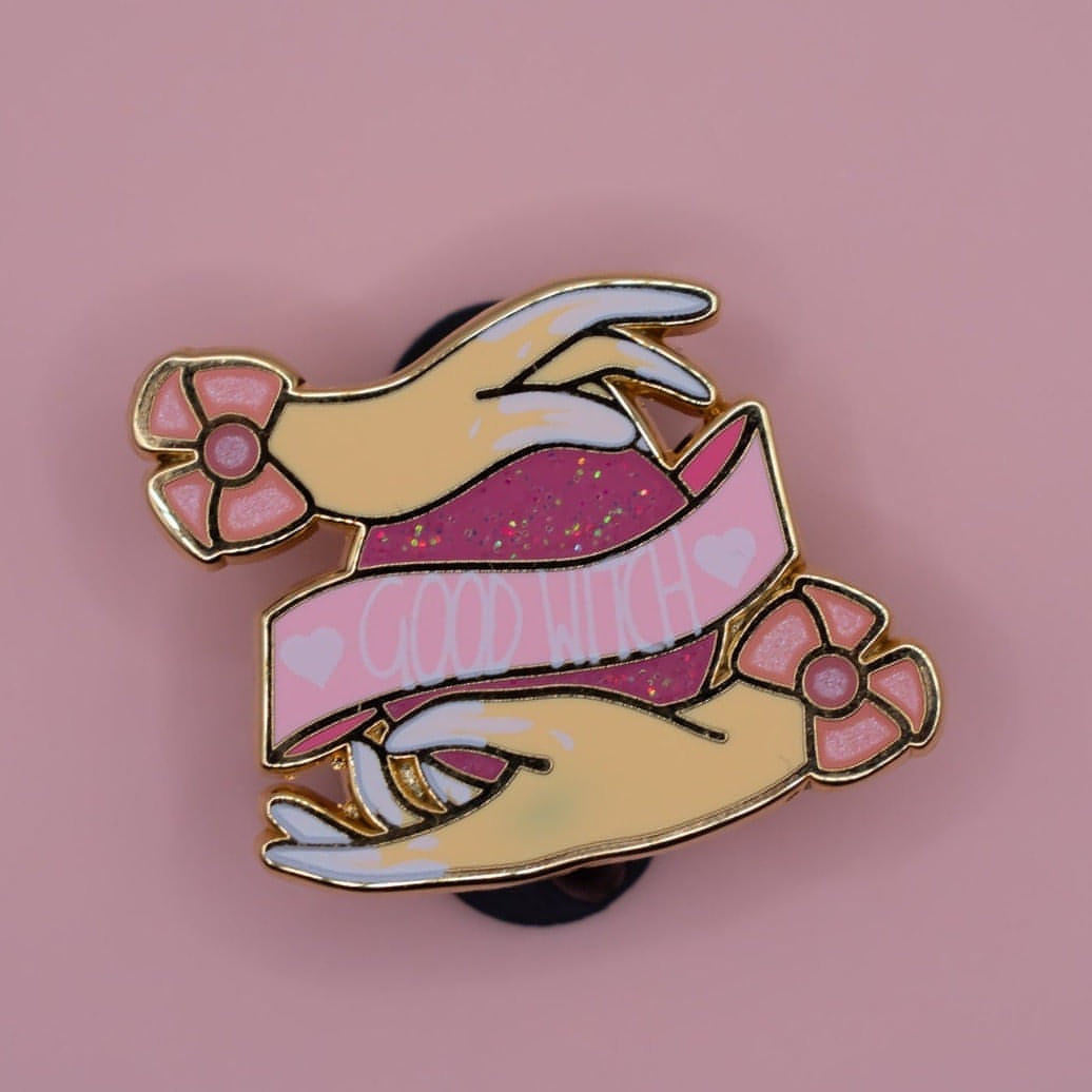 On a pink background is a hard enamel pin made of gold metal. In the centre is a hot pink orb with iridescent glitter and a banner ontop which says "good witch" in white capital letters. At the top and bottom of the orb is a hand with light tone skin. The tips of the fingers on each hand have white liquid screenprinted on. Each hand has a bow at the wrist which is light pink with a slightly darker pink centre. Each bow is pearl enamel to look shimmery and 3D.