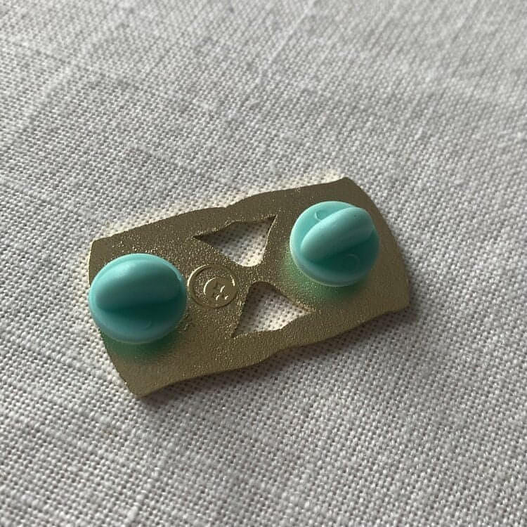 A photo of the back of the pin which shows the shop logo stamped in metal in the centre of the bottom half, and 2 round rubber backs - one at the top of the pin, one at the bottom.