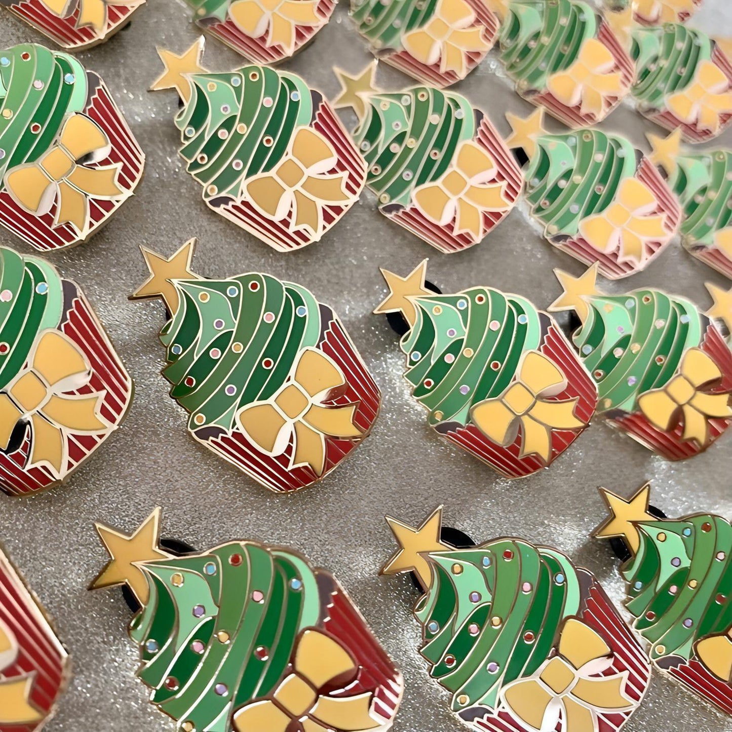 The image shows rows of many pins. The pins are in the shape of cupcakes with a yellow star at the top. Each cupcake has a red bottom, brown sponge, and the icing is different shades of green. There is a yellow bow on the red cup and there are multicoloured circles as sprinkles on the green enamel areas.