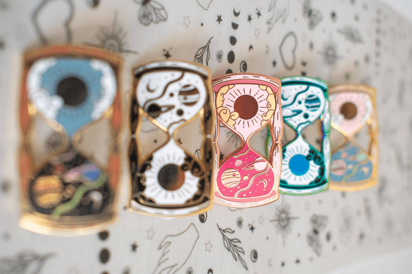 A photo showing five variations of the hourglass pin with different enamel colours and metal plating.