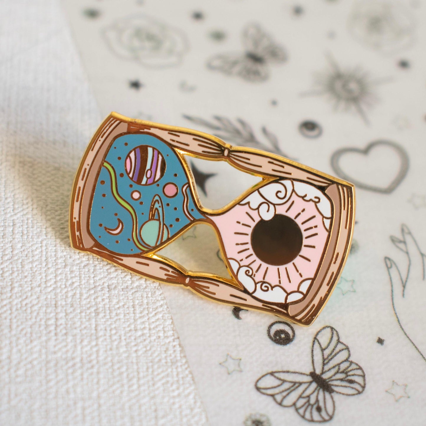 Pictured is an hourglass with a space scene and planets in the top half and a sun with some clouds in the bottom half. The hourglass is a mix of pastel pink, green, and blue enamel, and it is made from gold metal plating. It is hard enamel.