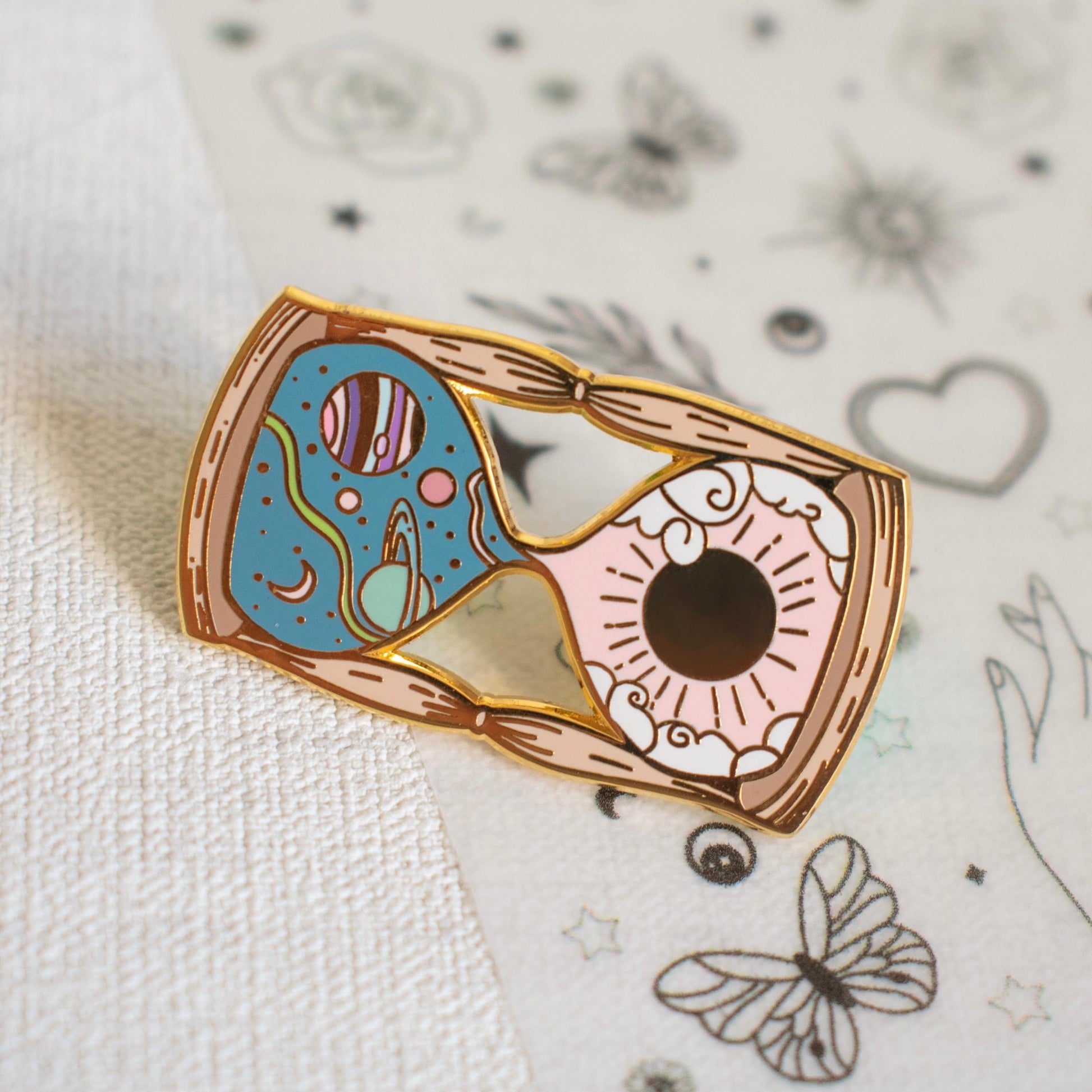 Pictured is an hourglass with a space scene and planets in the top half and a sun with some clouds in the bottom half. The hourglass is a mix of pastel pink, green, and blue enamel, and it is made from gold metal plating. It is hard enamel.