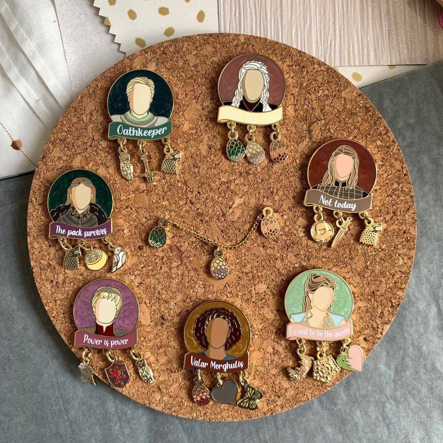 The picture shows all 7 Women of Westeros pins, each with three charms, a shimmery pearl swirl background, and the image of a character with a relevant quote in screenprint below.