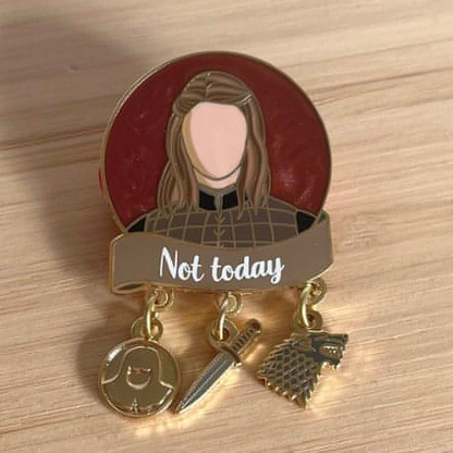 The photo shows a round pin with a dark red pearl swirl background which is shimmery. The head and shoulders of the character Arya is pictured in the centre of the circle in a black and brown shirt. Below is a brown banner with the quote "Not today" written in white screenprint. Below that are three charms which dangle by one ring. The charms are a Braavosi coin, a dagger, and the wolf sigil of House Stark.
