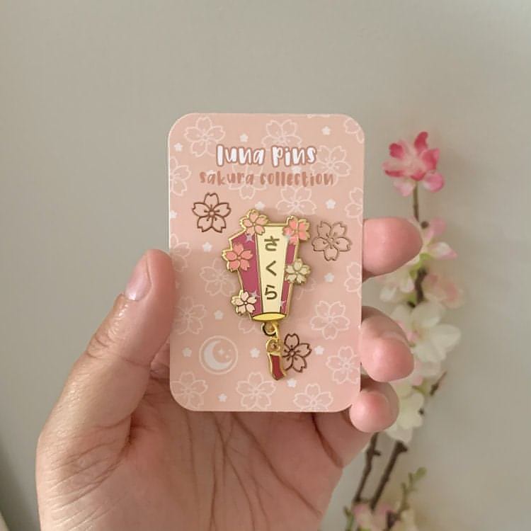 Someone is holding the pin on a special edition backing card. The card has a sakura pattern and rose gold foil effects.