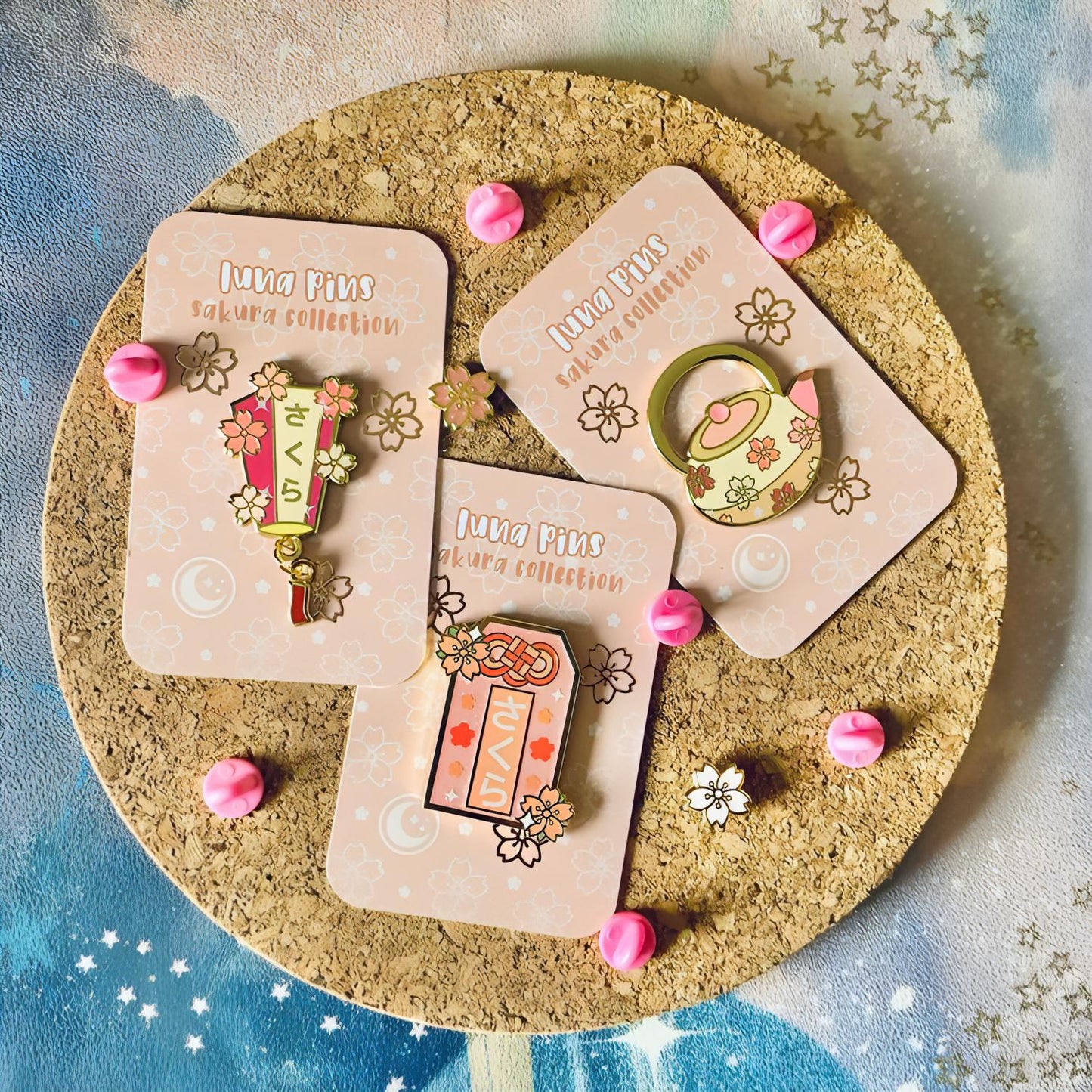 The pin is pictured with other pins in the sakura collection, including a teapot and a omamori charm. There are also miniature sakura flower pins.