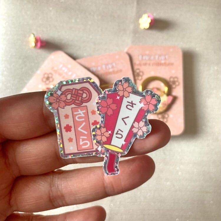 Someone holds two glitter stickers with the lantern design and the omamori design printed on.