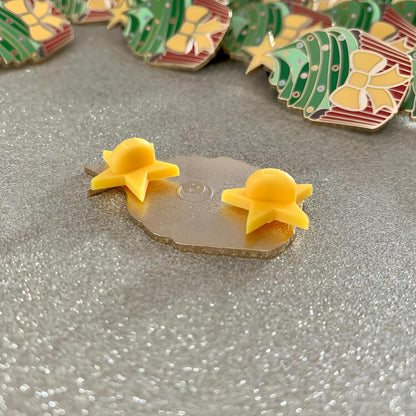 The image shows the back of the pin which features the shop logo and two yellow star shaped rubber backs at the top and bottom.