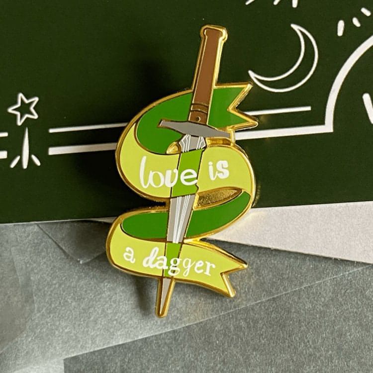 A grey dagger with a brown handle made of gold metal and hard enamel is pictured. The pin has a green enamel ribbon around the dagger. The phrase "love is a dagger" in a Loki TV series font is screenprinted in white on the ribbon.