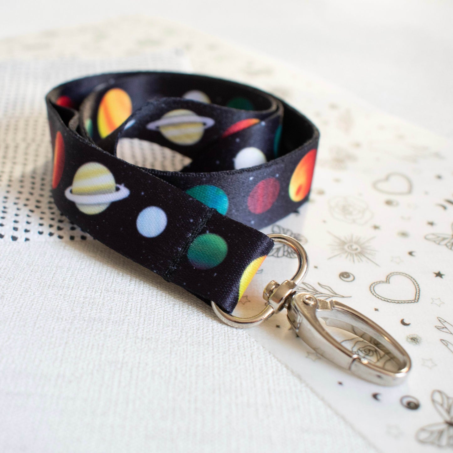  A black lanyard is pictured with full colour planetary pattern going up the length of it. Each planet is illustrated in full colour. The lanyard has an oval hook accessory at the end.
