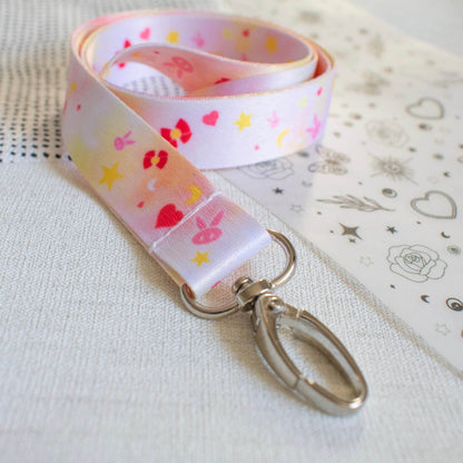 The image shows a pink and yellow pastel lanyard. The lanyard is fabric and has an anime pattern on it featuring bunny heads, hearts, bows, stars, and moons in different shades of yellow and pink. At the end of the lanyard is a silver oval hook.