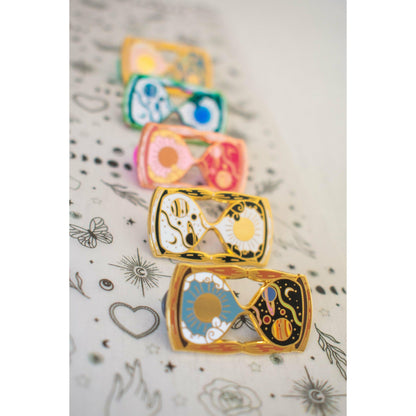 A photo showing five variations of the hourglass pin with different enamel colours and metal plating.