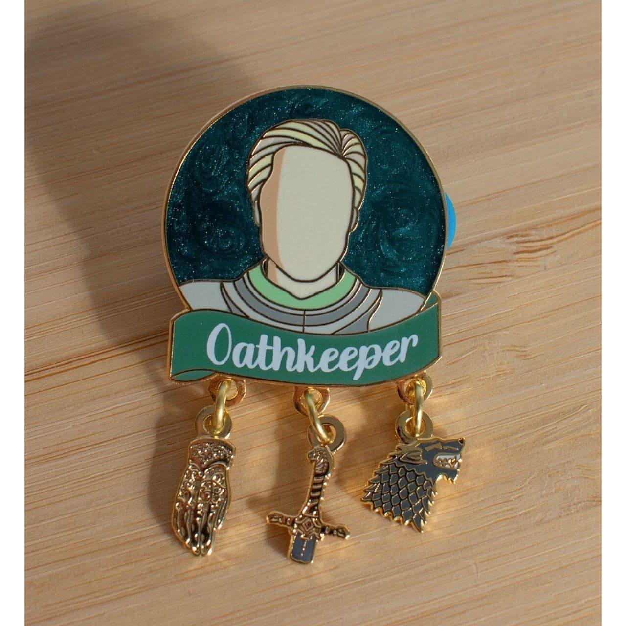 The photo shows a round pin with a dark blue pearl swirl background which is shimmery. The character Brienne is pictured in the centre of the circle in her Game of Thrones armour. Below is a green banner with the quote "Oathkeeper" in white screenprint. Below that are three charms which dangle by one ring. The charms are Jaime's golden hand, the hilt of her sword, and the Stark wolf sigil.