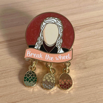 The photo shows a round pin with a pinky red pearl swirl background which is shimmery. The head and shoulders of the character Daenerys is pictured in the centre of the circle in a black dress with a dark red neck collar. Below is a pale orange banner with the quote "Break the wheel" in white screenprint. Below that are three charms which dangle by one ring. The charms are each of her dragon eggs.