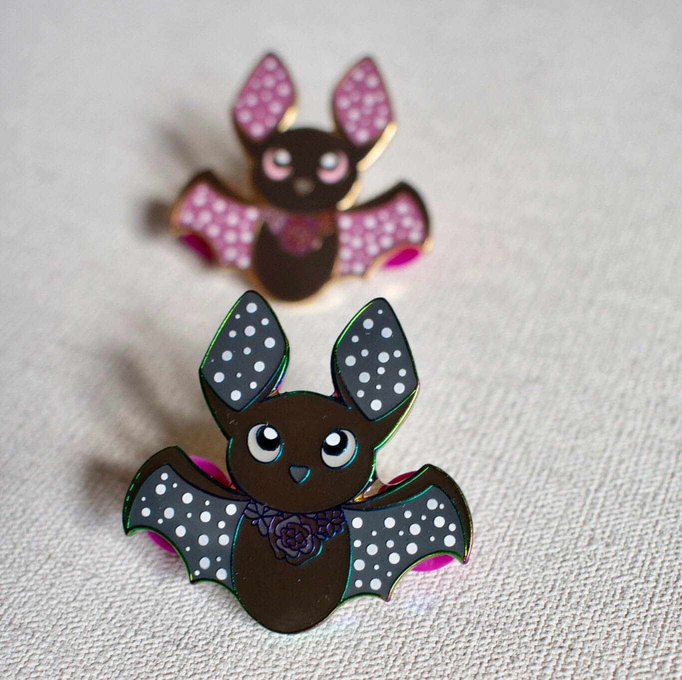 At the front of the image is the bat pin with rainbow metal plating. The bat is black with grey wings and white dots on its ears and wings. At the back of the image is the bat pin with purple ears and wings and a black body.