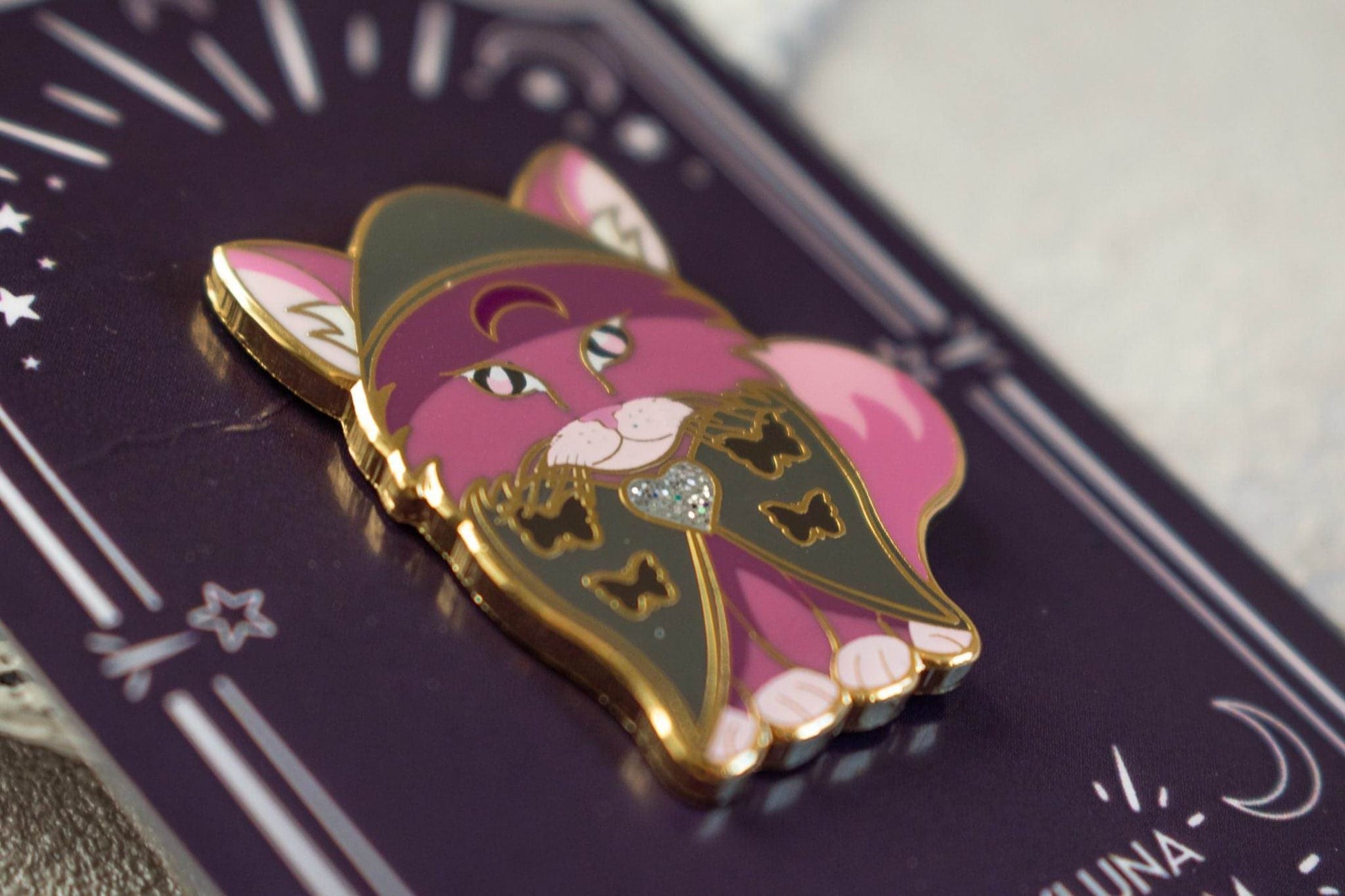 A close up shot of the cat pin shows the small bits of glitter in the hart brooch and also the quality of the screenprinted face and eyes. The cat has darker grey enamel butterflies on its cape.
