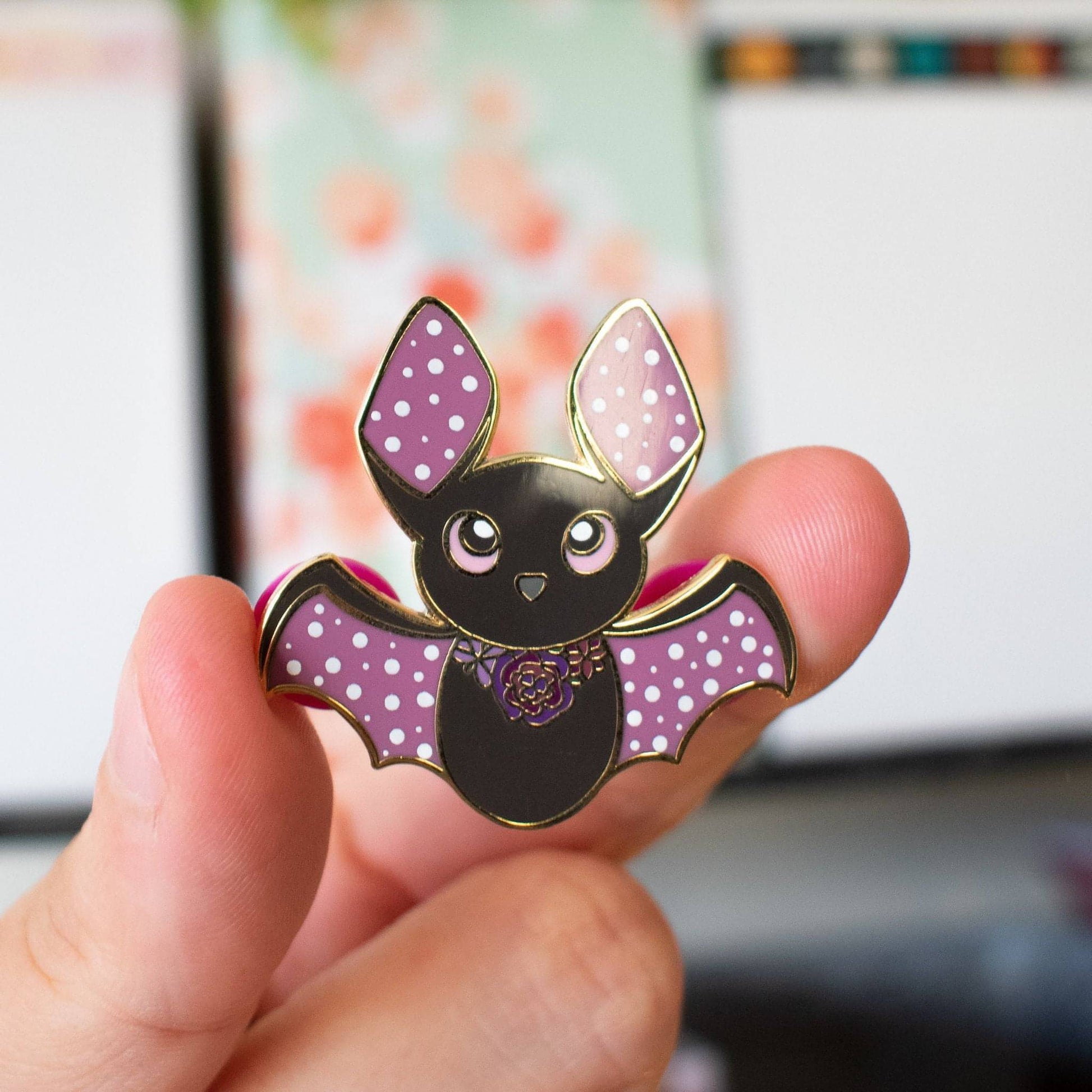 A black hard enamel bat pin with purple ears and wings. It has white dots on its ears and wings and a floral collar.