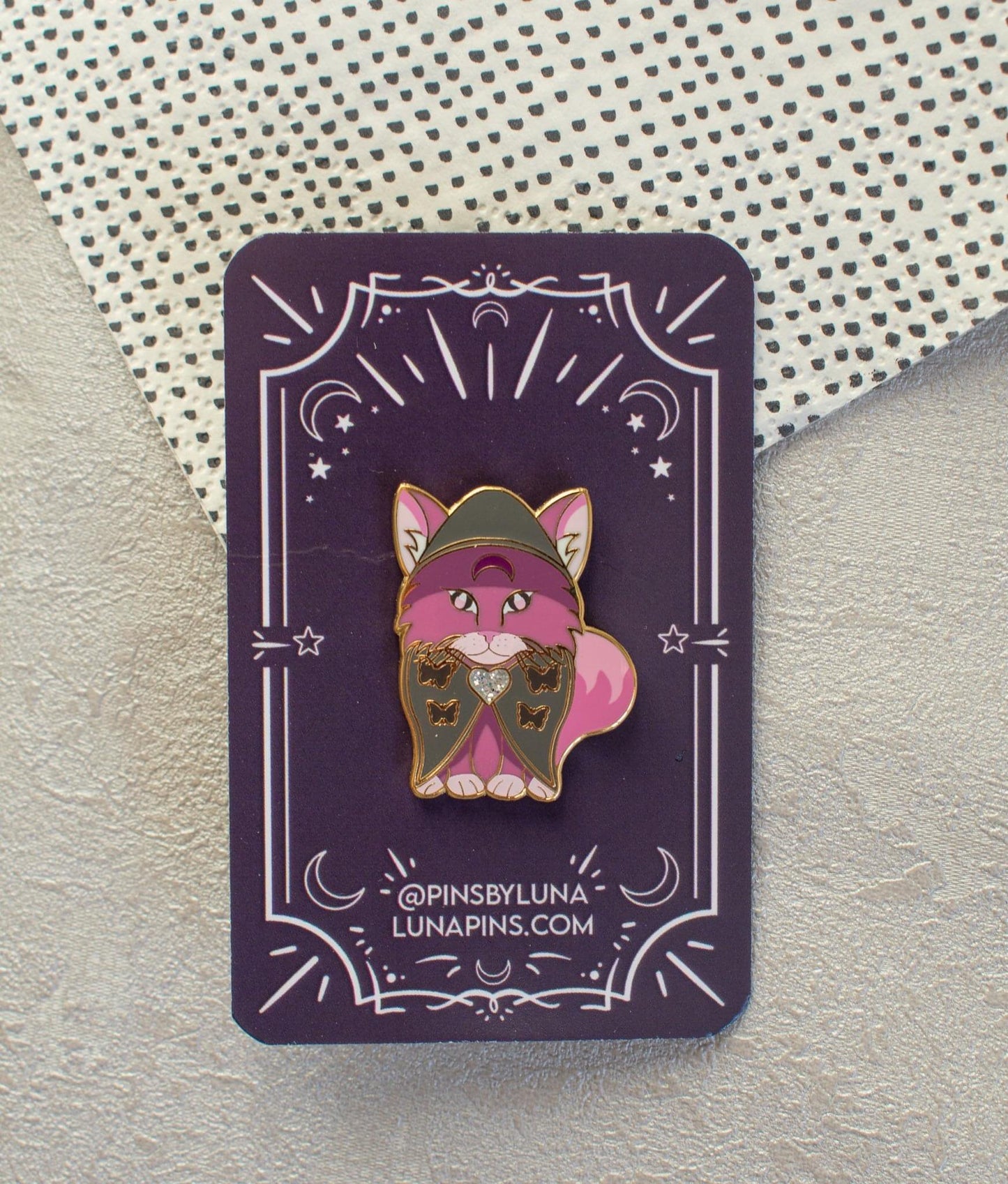 A head-on image of a purple cat enamel pin. The cat wears a dark grey cape with a hood and a silver glitter brooch in the centre. The cats paws and inner ears are a lighter purple and it has an upside down crescent moon in dark pin on its forehead.