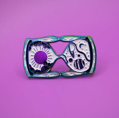 The image shows the rainbow metal plated black and white hourglass pin on its side on a bright purple background.