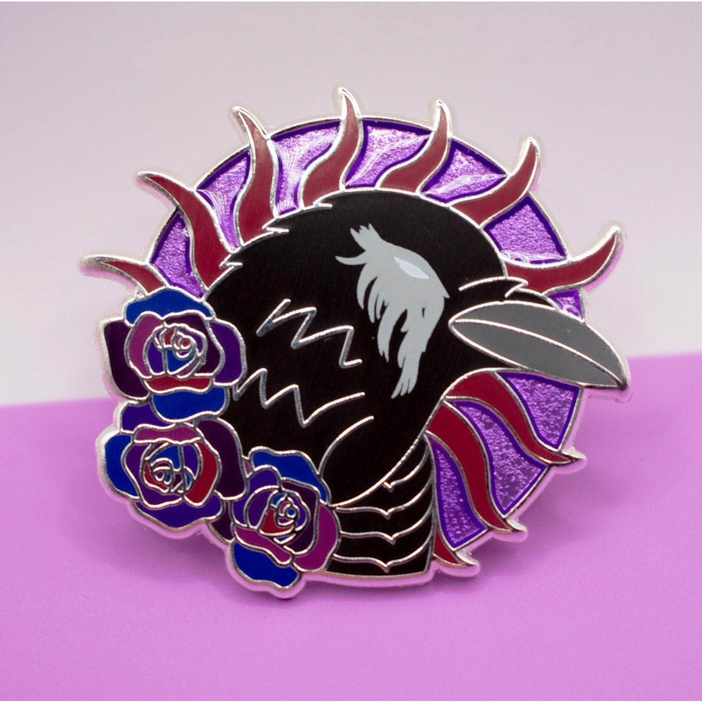 In the photo is a silver metal pin featuring a black crow looking right. The crow has a grey beak and a white eye with grey feathers around it. The crow is on a round purple background with violet spokes going around it. The round purple background is transluscent enamel to look 3D.  Beneath the crow at its neck are three roses in various shades of purple.