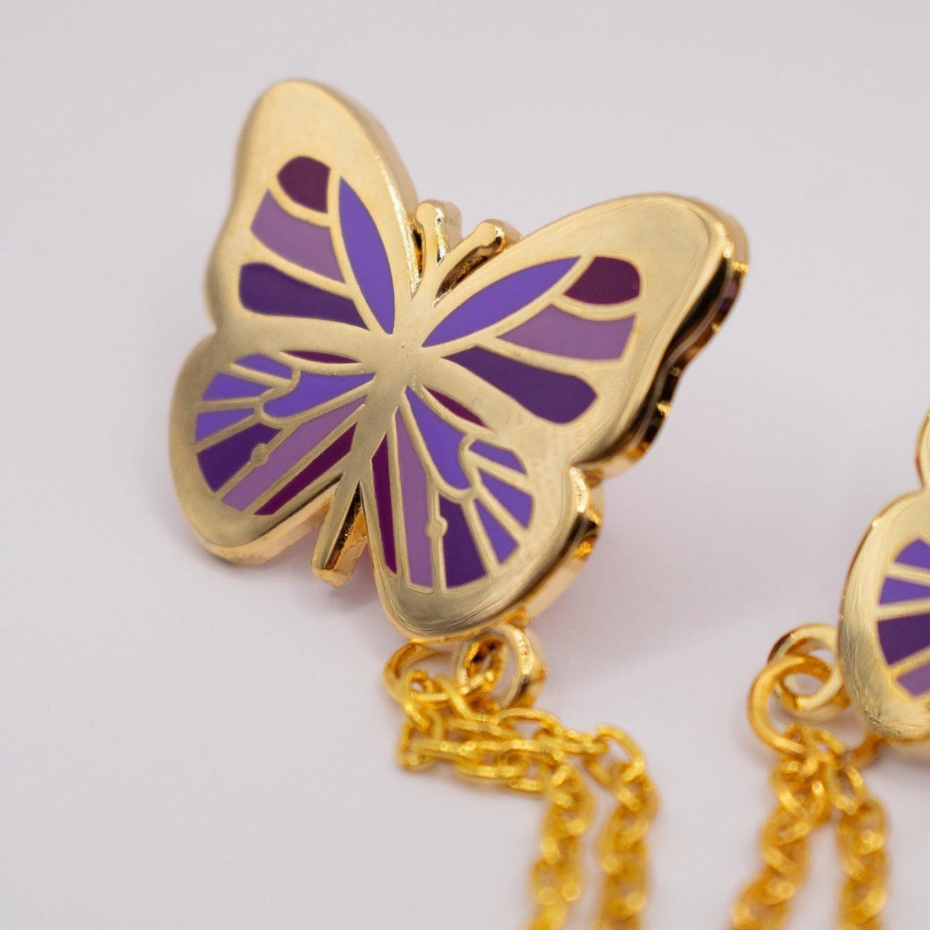 Pictured is a close up shot of one of the purple butterlies and the chain attaching it to the other butterfly. The butterfly wings contain various shades of purple, the outside metal is a shiny gold, and the chain is also gold.