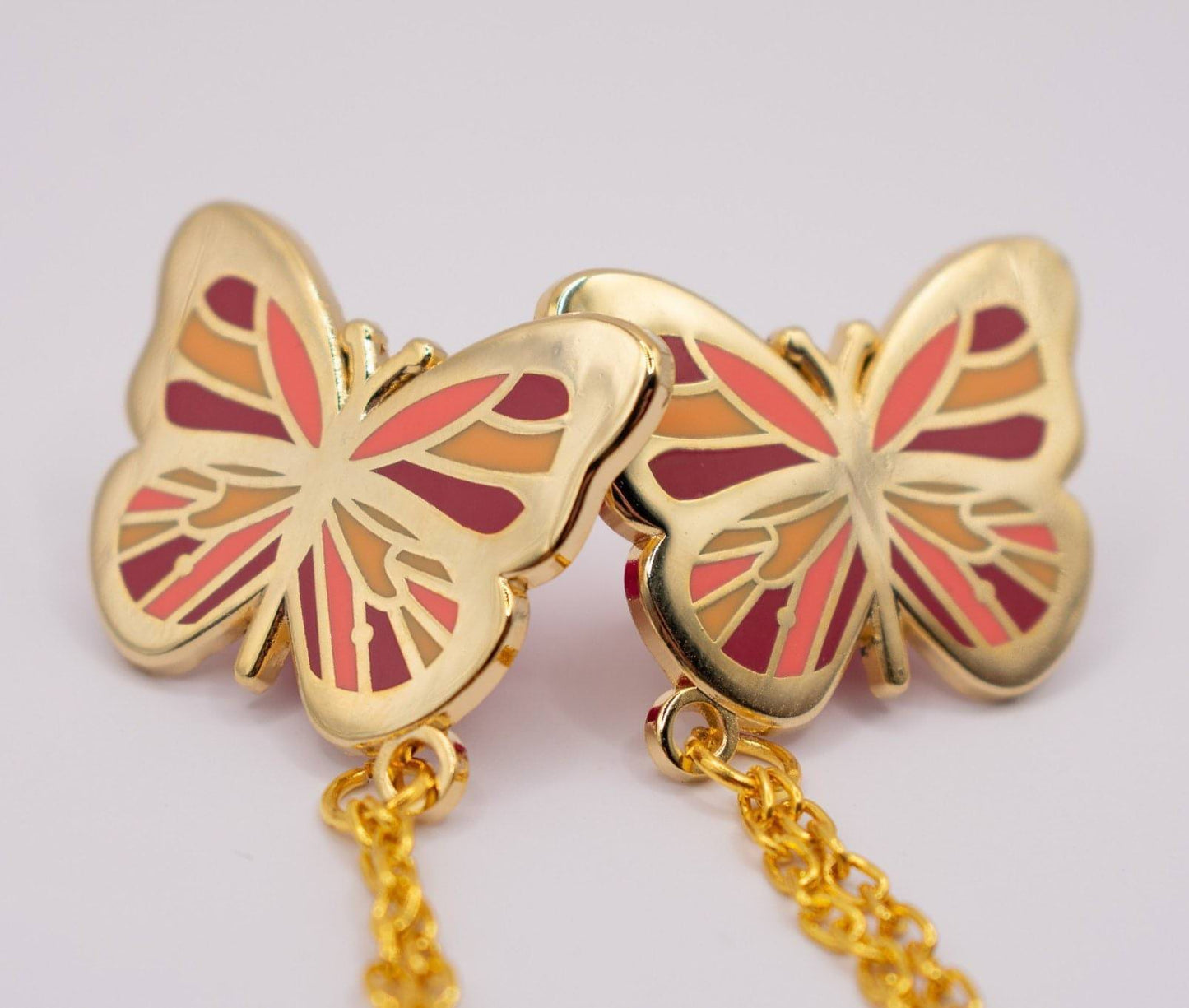 This picture shows the butterfly collar pin (which is two butterfly pins connected by chains). In this image is the pink variant, which means the wing colours are various shares of pink and orange. The metal of the pin is a shiny gold.